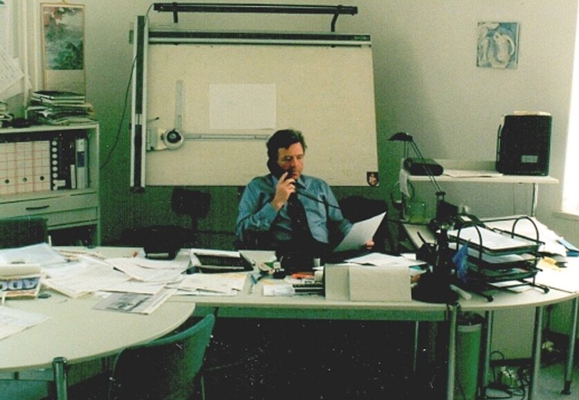 A man in an office sits at a cluttered desk, reading papers and holding a pen to his mouth. Behind him is a large drafting board, shelves with files, and a window letting in natural light.